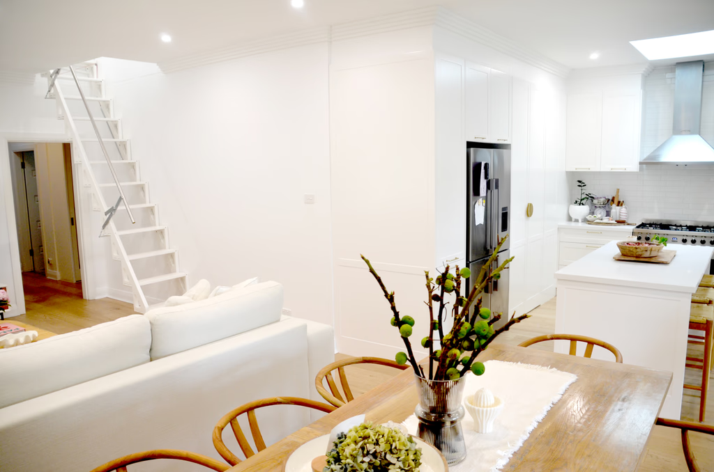 Slim white space-saving folding staircase with timber-edged treads rising against the wall of a bright open-plan Sydney home